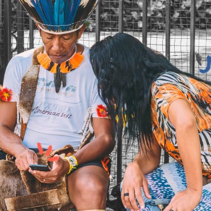 Linklado: Um homem indígena utiliza um celular. Uma mulher olha para a tela do aparelho. Atrás deles, grades, uma tela de proteção e uma mochila em preto e branco.