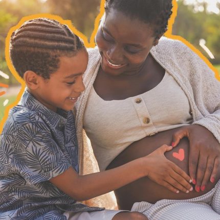 Uma mulher negra gestante está sentada no gramado, acompanhada do filho, também negro. O menino e a mulher fazem um coração com as mãos na barriga da mãe.