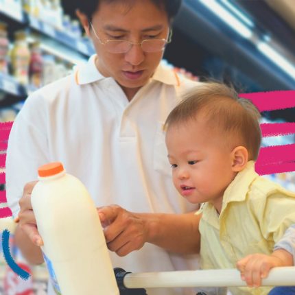 Um homem asiático está com uma garrafa de leite nas mãos. Ao lado, uma criança asiática, em cima de um carrinho de supermercado observa o objeto; nas prateleiras outros produtos lácteos da seção.