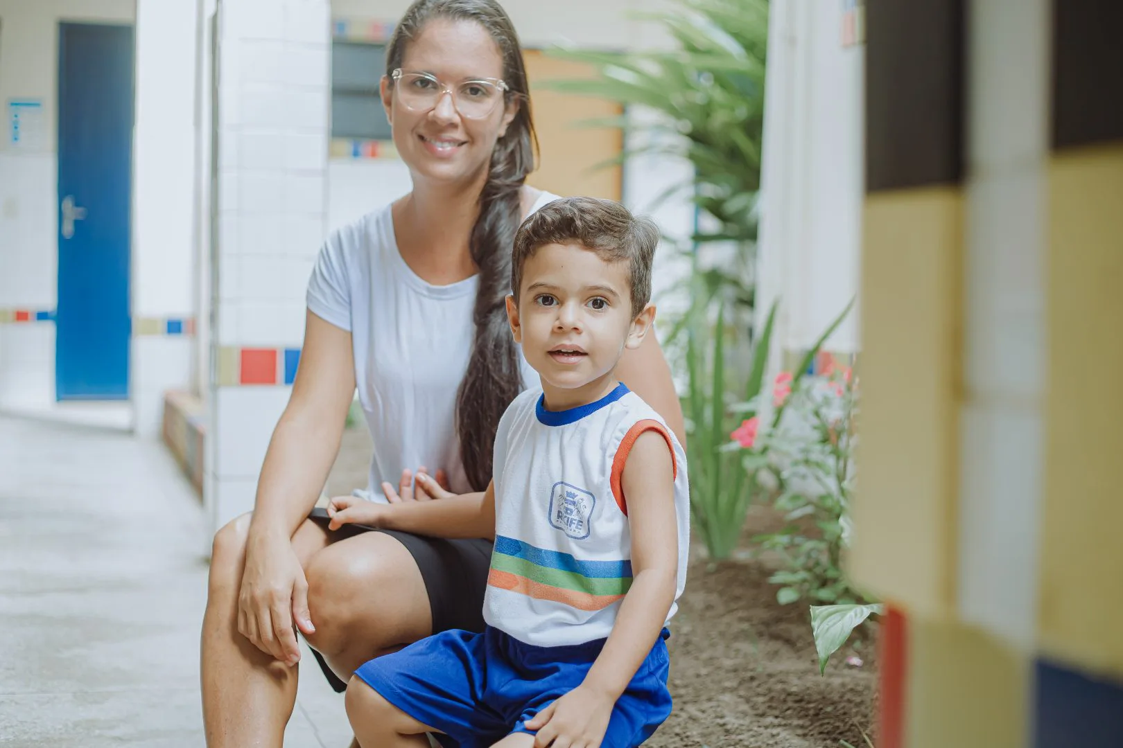 Uma mulher de óculos e cabelos longos e lisos, vestindo camiseta branca e saia preta, está sentada ao lado de um menino que veste uniforme escolar Ao fundo, o ambiente de uma escola, com o corredor que dá acesso a salas de aula e algumas plantas.