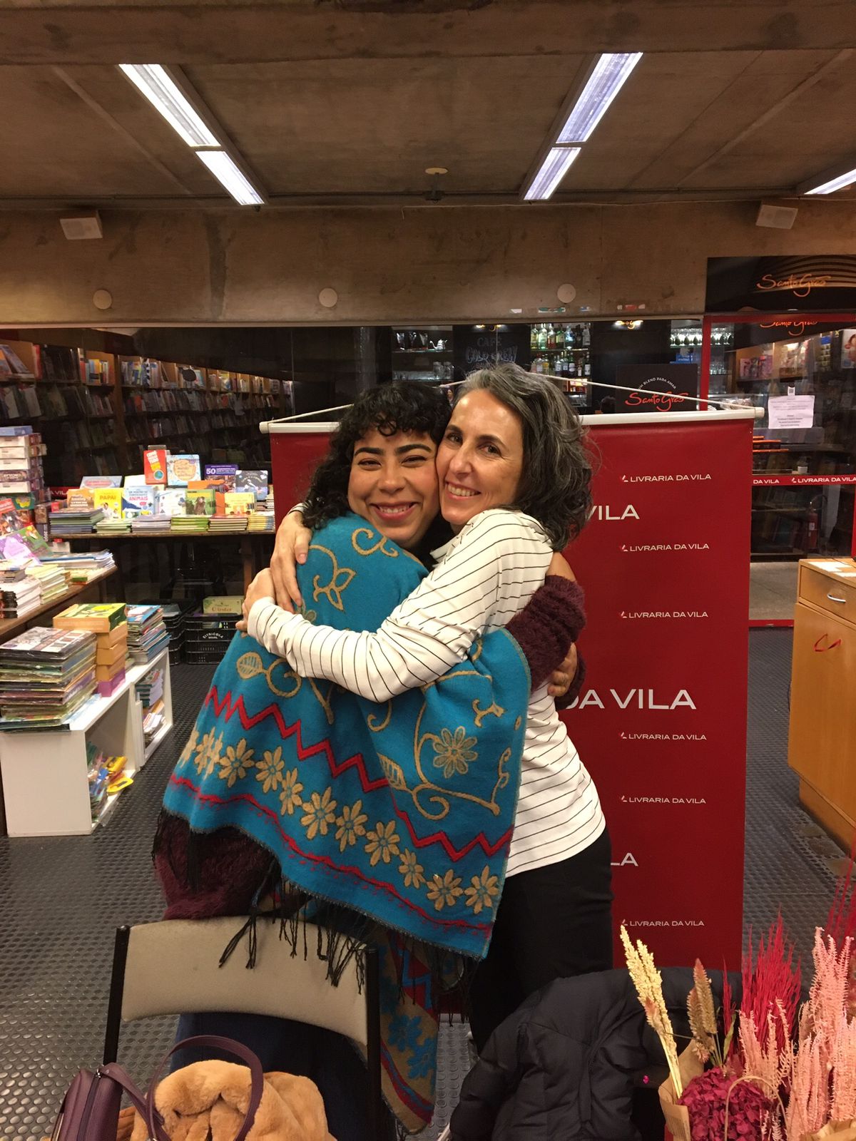 Foto das autoras Angélica Kalil e Amma, do livro "Amigas que se encontraram na história". São duas mulheres brancas se abraçando em uma livraria.
