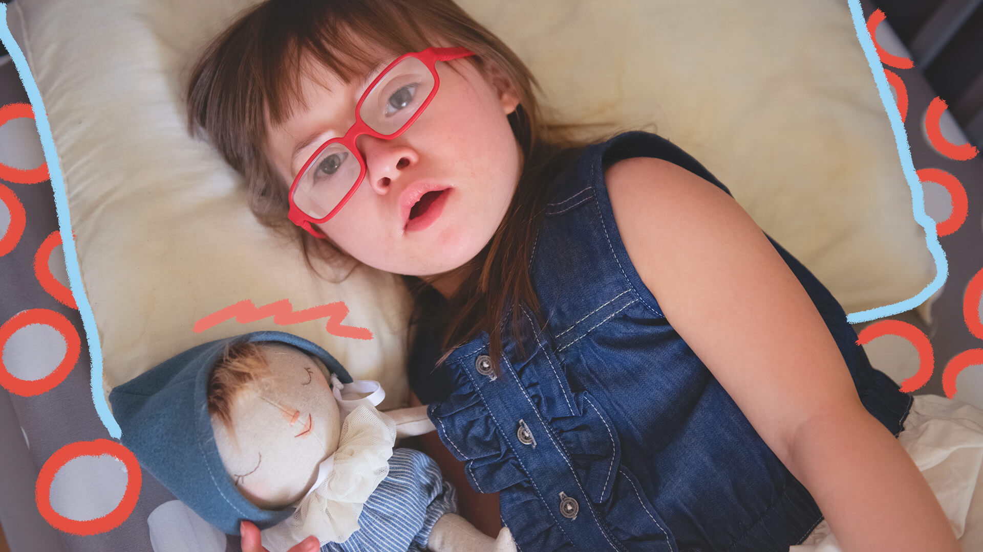 Foto de uma menina que tem síndrome de Down deitada com sua boneca. Ela usa roupa jeans e a boneca usa azul