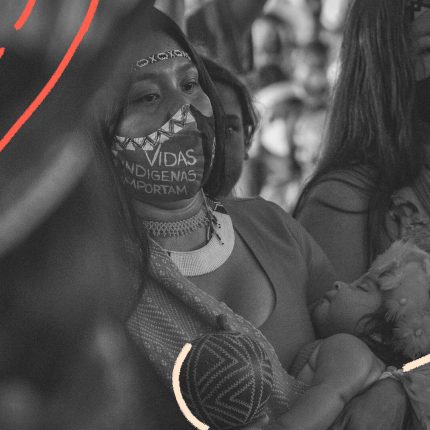 Foto em preto e branco de mulheres indígenas. A mulher em primeiro plano usa máscara e carrega no colo um bebê. A matéria é sobre Mortes violentas de crianças no AM