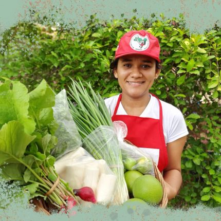 Aline Oliveira, da juventude do MST, está com um cesto de verduras e legumes na mão. Ela é uma mulher de pele parda, sorrindo, vestindo uma camisa branca, um avental vermelho e um boné do MST. Ao fundo, uma árvore, e ao redor, uma intervenção de fundo verde com ilustração de folhas.