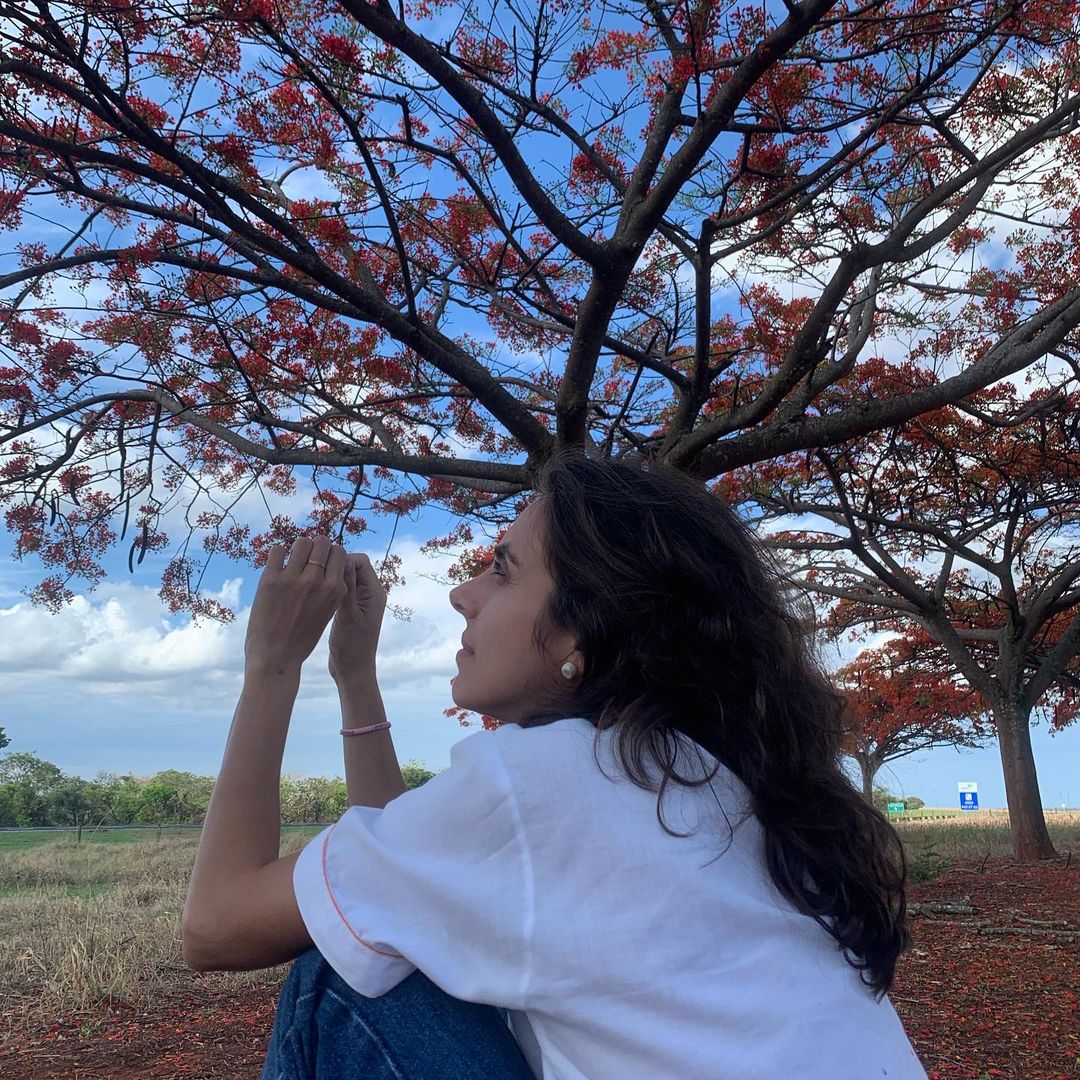 Giovanna Nader, vestindo camiseta branca e calça jeans, com cabelos escuros e soltos, está embaixo de uma árvore, olhando para o céu.