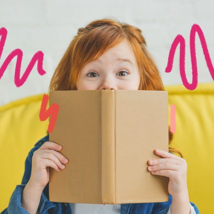 Melhores livros de 2022: foto de uma menina ruiva que segura um livro de capa lisa na cor bege com as duas mãos, em frente à boca. Ela está sentada num sofá amarelo.