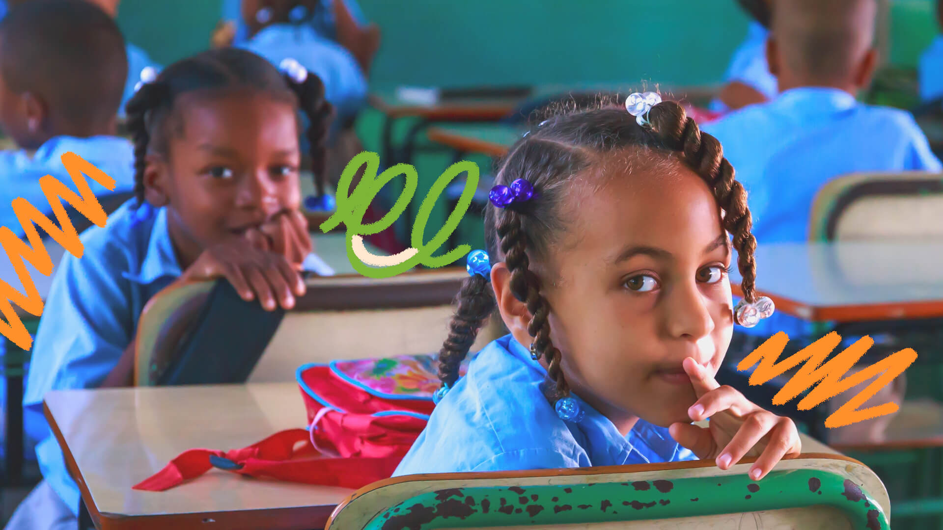 Foto de crianças em uma sala de aula. Elas vestem uniforme azul e duas meninas olham para a foto