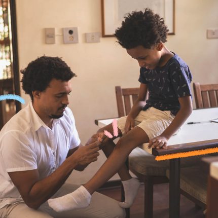 Um pai está agachado ajudando o filho, sentado na mesa, a calçar o sapato. Ambos são negros. O homem usa roupas claras e o menino usa camiseta azul marinho. O texto é sobre paternidade.