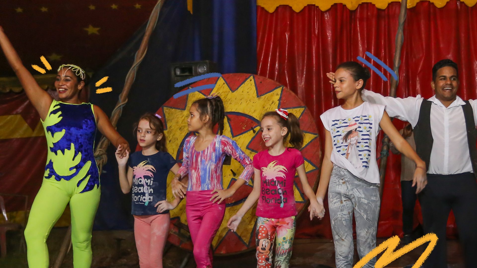 Foto de um grupo de crianças se apresentando no circo. Todas vestem roupas coloridas.