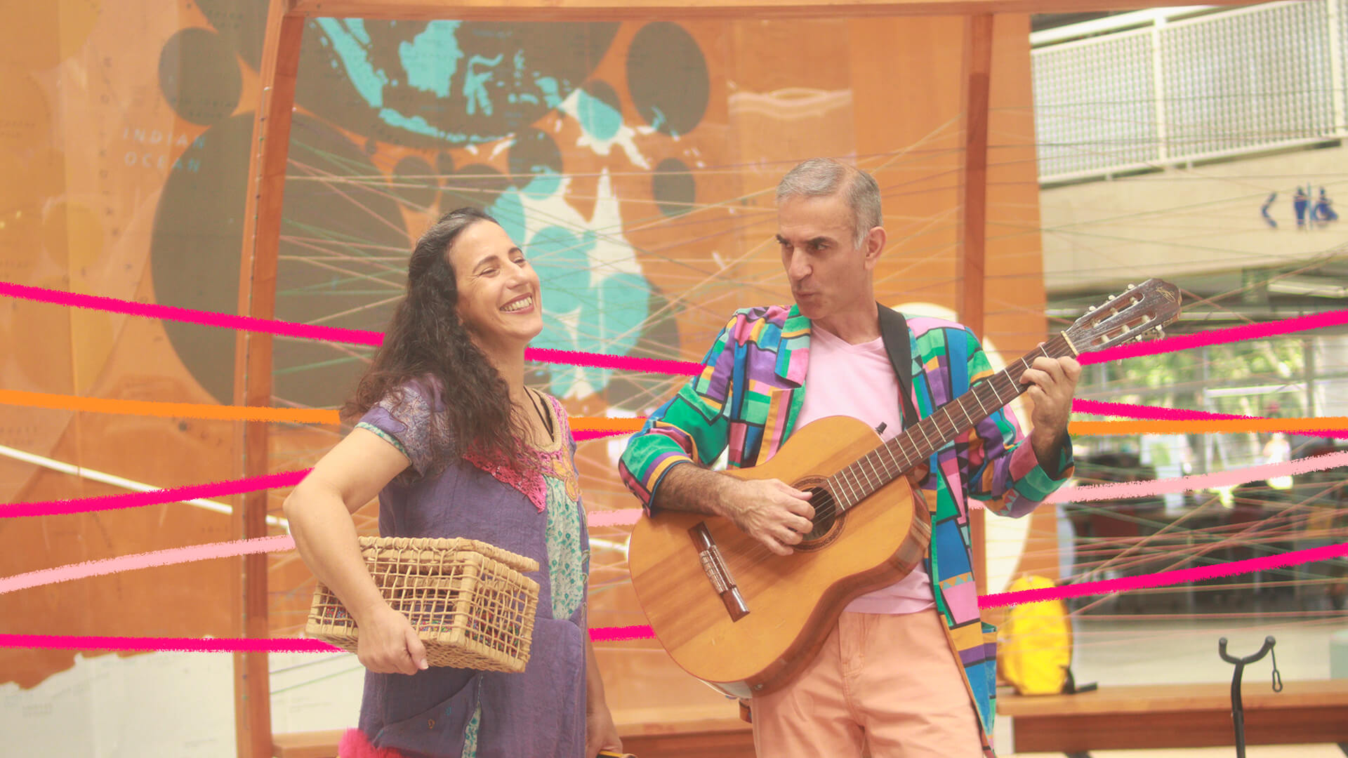 Uma mulher e um homem vestem roupas coloridas. Ela segura uma caixa e ele um violão. Eles são da Singela Trupe, companhia de teatro que apresenta Machado de Assis para as crianças