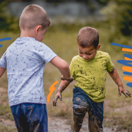 Foto de dois meninos brincando na lama. As roupas são coloridas e estão sujas de barro.