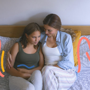 Mãe e filha adolescente estão sentadas conversando. A garota está com os braços cercando a região da barriga. A imagem ilustra uma matéria sobre primeira menstruação.
