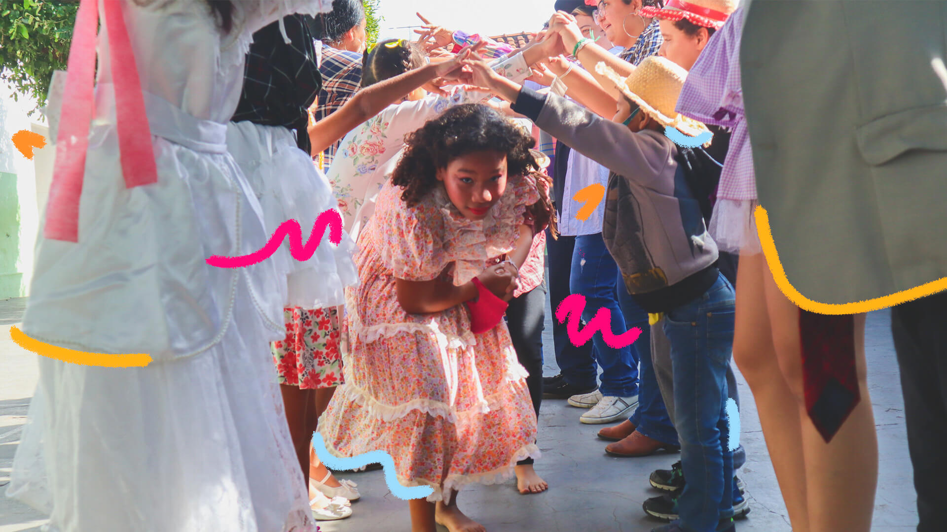 Uma menina com vestido estampado e colorido, típico de festa junina, passa debaixo de um túnel formado por pares de mãos dadas. A matéria é sobre crianças no São João.