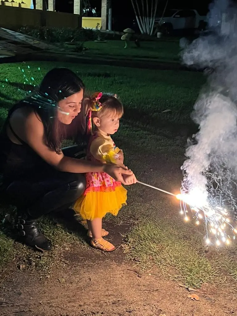 Uma mulher está ajoelhada próxima de uma menina vestida de festa junina soltando fogos