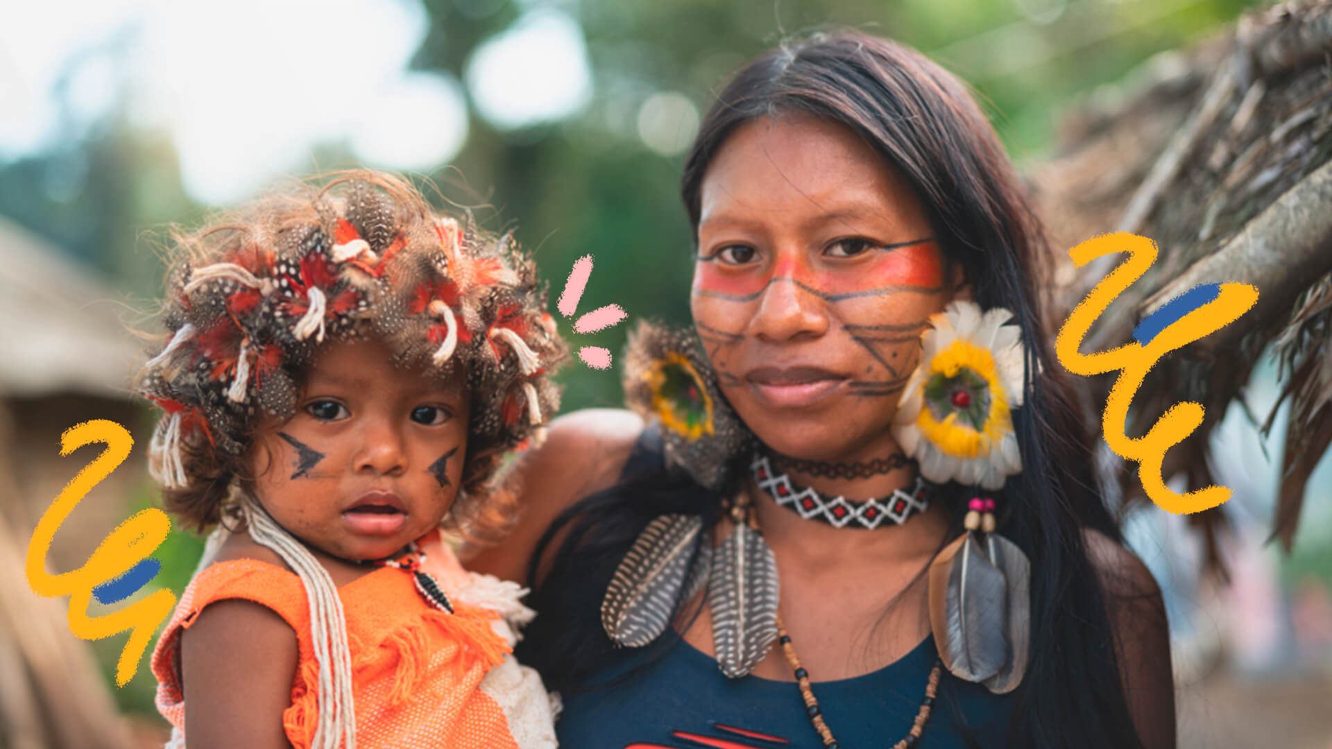 Imagem com mulher indígena segurando no colo uma criança indígena. Ambos estão com os rostos pintados e usam acessórios indígenas.