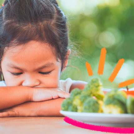 Menina branca d ecabelos castanhos repousa os braços sobre a mesa ao recusar um prato de brócolis