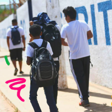 Um grupo de quatro jovens caminham por uma calçada, em paralelo a um muro. Eles usam uniforme escolar e três deles carregam mochilas nas costas