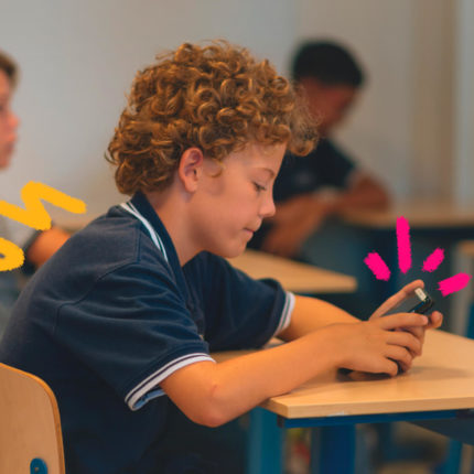 Imagem mostra um menino branco de cabelos cacheados vestindo camisa azul e usando um celular dentro de uma sala de aula, sentado na carteira.