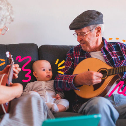Na imagem, um homem idoso e um bebê, avô e neto, estão sentados um ao lado do outro em um sofá. O avô toca um instrumento de cordas e sorri para o neto. Ambos possuem pele branca.