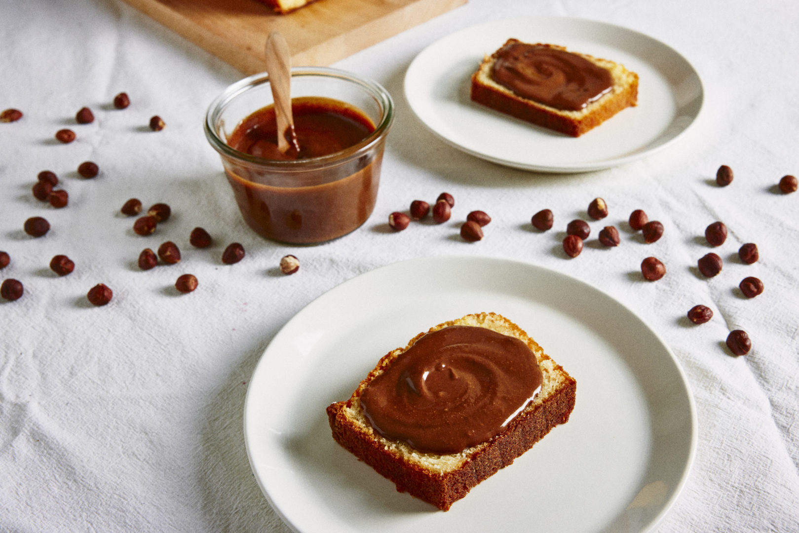 Receita de nutella caseira: o creme de avelã marrom está em um copo transparente e em cima de uma fatia de bolo em um prato branco sobre a mesa soberta de gotas de chocolate.