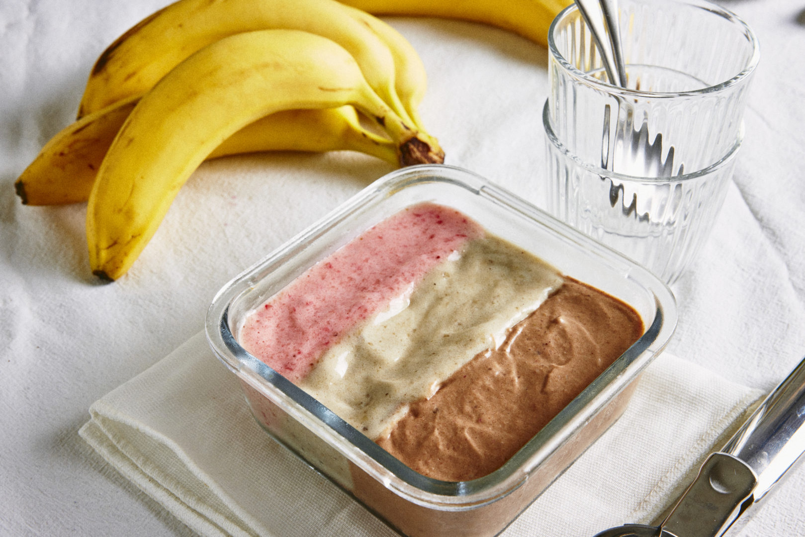 Receita de sorvete napolitano: em cima da mesa há um cacho de bananas amarelas ao lado de uma taça trnasparente e um pote com o sorvete de três cores - rosa, branco e marrom. 