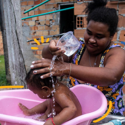 Mulher dá banho em bebê jogando água de uma vasilha de plástico em uma banheira no quintal de casa. Imagem ilustra a capa da matéria especial sobre saneamento e crianças.