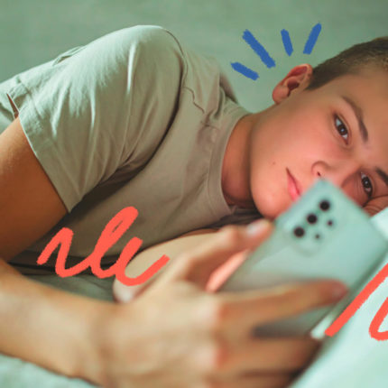 Um menino de pele clara está deitado na cama e olha para a tela de um celular. A imagem ilustra matéria sobre notificações no celular.