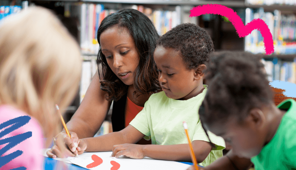 Imagem para matéria sobre educação antirracista mostra uma professora negra sentada aolado de dois alunos, um menino e uam menina negros, que escrevem com lápis em um papel.