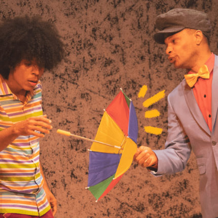 Imagem mostra cena do espetáculo musical Bento Batuca, em que um ator negro de camisa listrada segura um guarda-chuva colorido de frevo, equanto outro ator negro usa chapéu, terno e gravata borboleta.