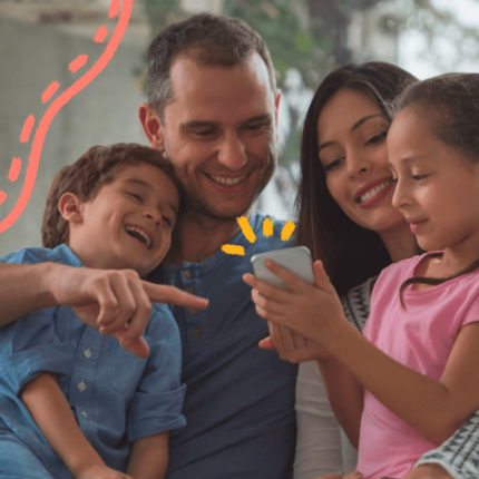 Foto de um casal com os dois filhos olhando para um celular. Quem segura o aparelho é a menina, que usa camiseta rosa.