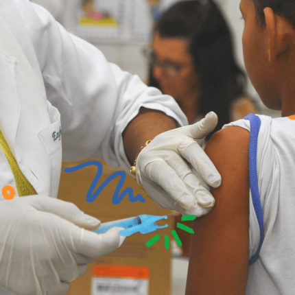 imagem ilustra uma nota sobre a vacina contra a dengue para crianças e mostra um menino de costas sendo vacinadado po rum aenfemeira, vestida de branco.