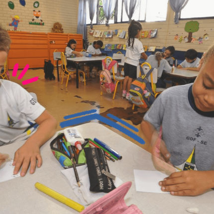 Imagem ilustra a matéria sobre a pesquisa educação infantil mostra uma sala de aula e duas crianças negras com uniforme, sentadas fazendo uma tarefa com lápis e papel.