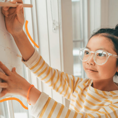 Imagem de capa de matéria sobre o ano bissexto mostra uma menina com traços asiáticos, usando óculos e cabelos presos. Ela escreve com um acaneta em um calendário fixado na parede.