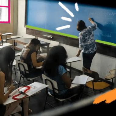 Três meninas estão sentadas em carteiras na sala de aula enquanto a professora escreve na lousa de cor azul.