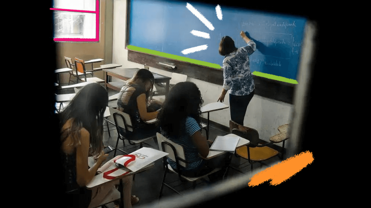 Três meninas estão sentadas em carteiras na sala de aula enquanto a professora escreve na lousa de cor azul.