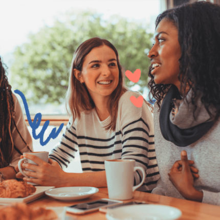 Um grupo de mulheres conversando e tomando café. Duas são morenas e a do meio é loira. As louças são brancas e tem um croissant na mesa.