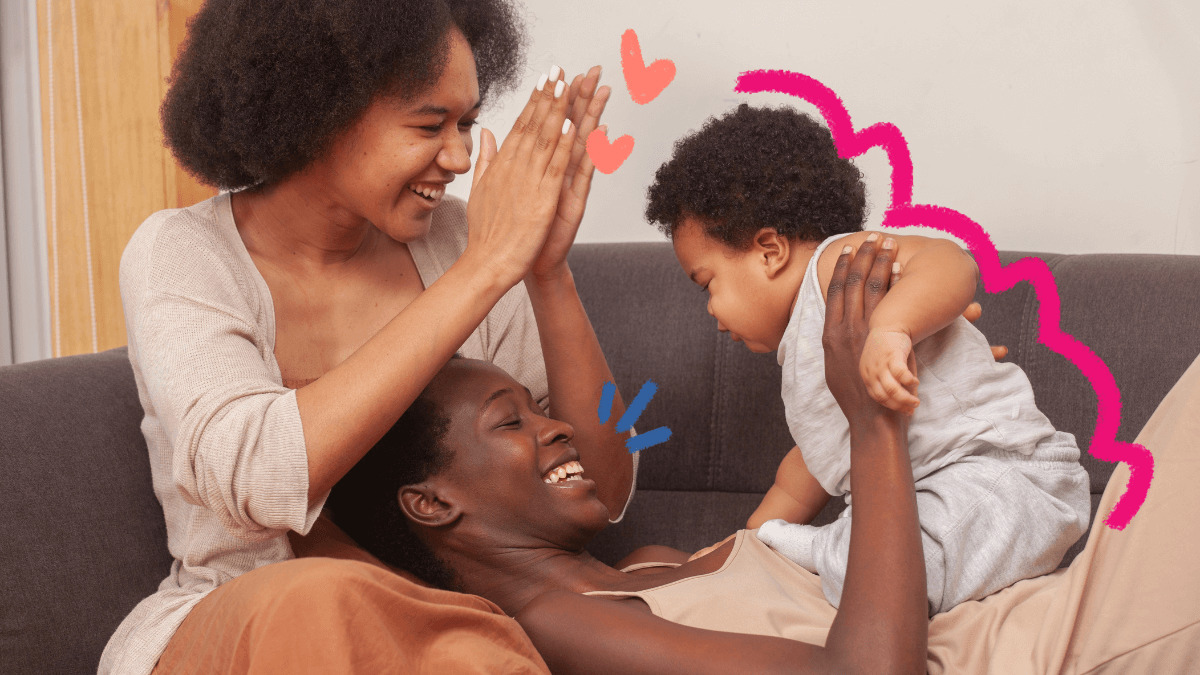 Duas mulheres brincam com um bebê no sofá. As três pessoas são negras e usam roupas claras.