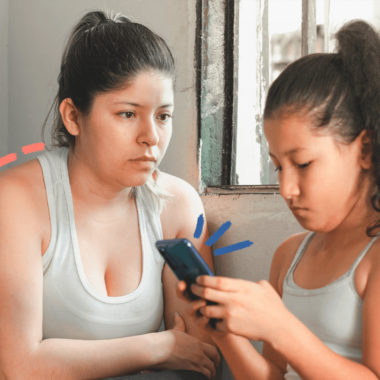 Uma mulher olha para uma menina enquanto ela usa o celular. Ambas têm cabelo escuro e usam regatas brancas. A matéria fala sobre como as mães que tentam equilibrar a relação telas e crianças estão sobrecarregadas.