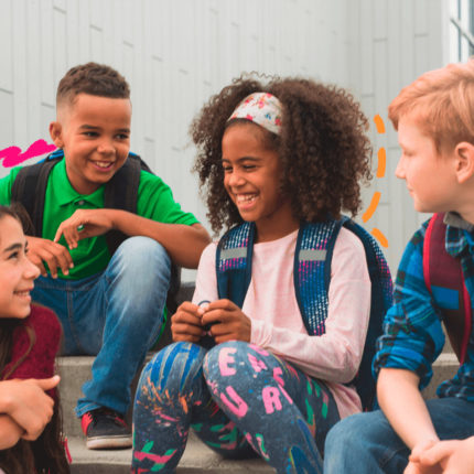 Um grupo de crianças de diferentes etnias e usando roupas coloridas estão sentadas e conversam entre si. A matéria traz os dados de um relatório da OCDE apontou que as competências socioemocionais favorecem a aprendizagem, o bem-estar e a saúde mental dos estudantes.