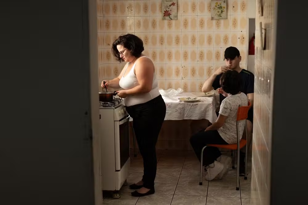 imagem da matéria de sobrecarga materna mostra uma mulher branca, de cabelos curtos e óculos preparando a comida em um acozinha e ao fundo os filhos sentados ä mesa.