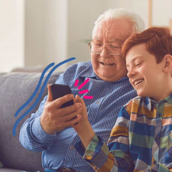 Um homem idoso de cabelos brancos e camisa azul divide com um menino ruivo de camiseta listrada a tela de um celular. Os dois estão sentados num sofá cinza. A matéria fala sobre a resolução do Conanda e a proteção de crianças no ambiente digital.