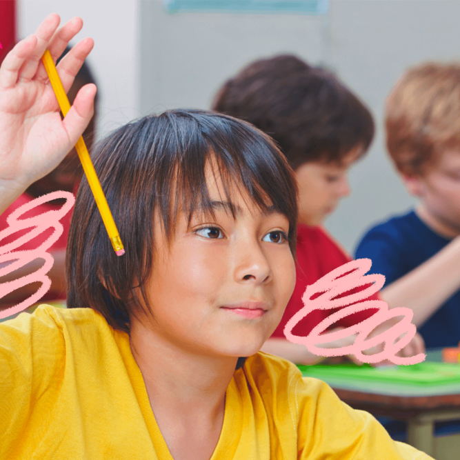 Um menino de cabelo escuro e camiseta amarela levanta a mão que segura um lápis amarelo para fazer uma pergunta na sala de aula. Outras crianças com roupas coloridas estão sentadas em suas respectivas carteiras.