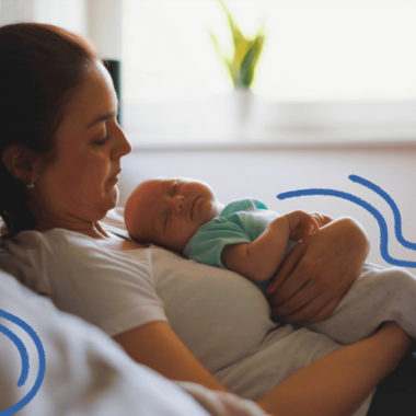 imagem para matéria sobre maternidade e vínculo mostra uma mulher branca, usando camisa branca e jeans sentada em um sofá de uma sala e segurando no colo um bebê recém nascido.