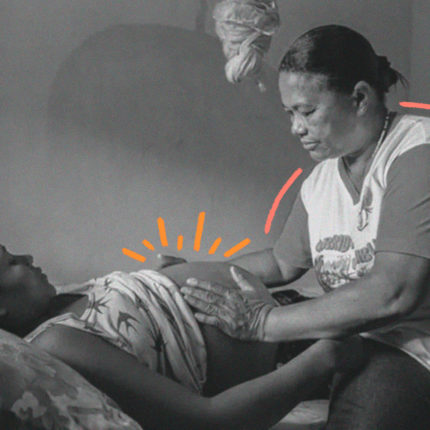 imagem para matéria sobre parteiras tradicionais mostra uma mulher mais velha sentada em uma cama tocando na barriga de uma mulher grávida que está deitada.