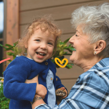 imagem de capa para matéria sobre avós e netos mostra uma mulher branca, idosa, de cabelos brancos, segurando no colo uma menina branca, d ecabelos cacheados e sorridente