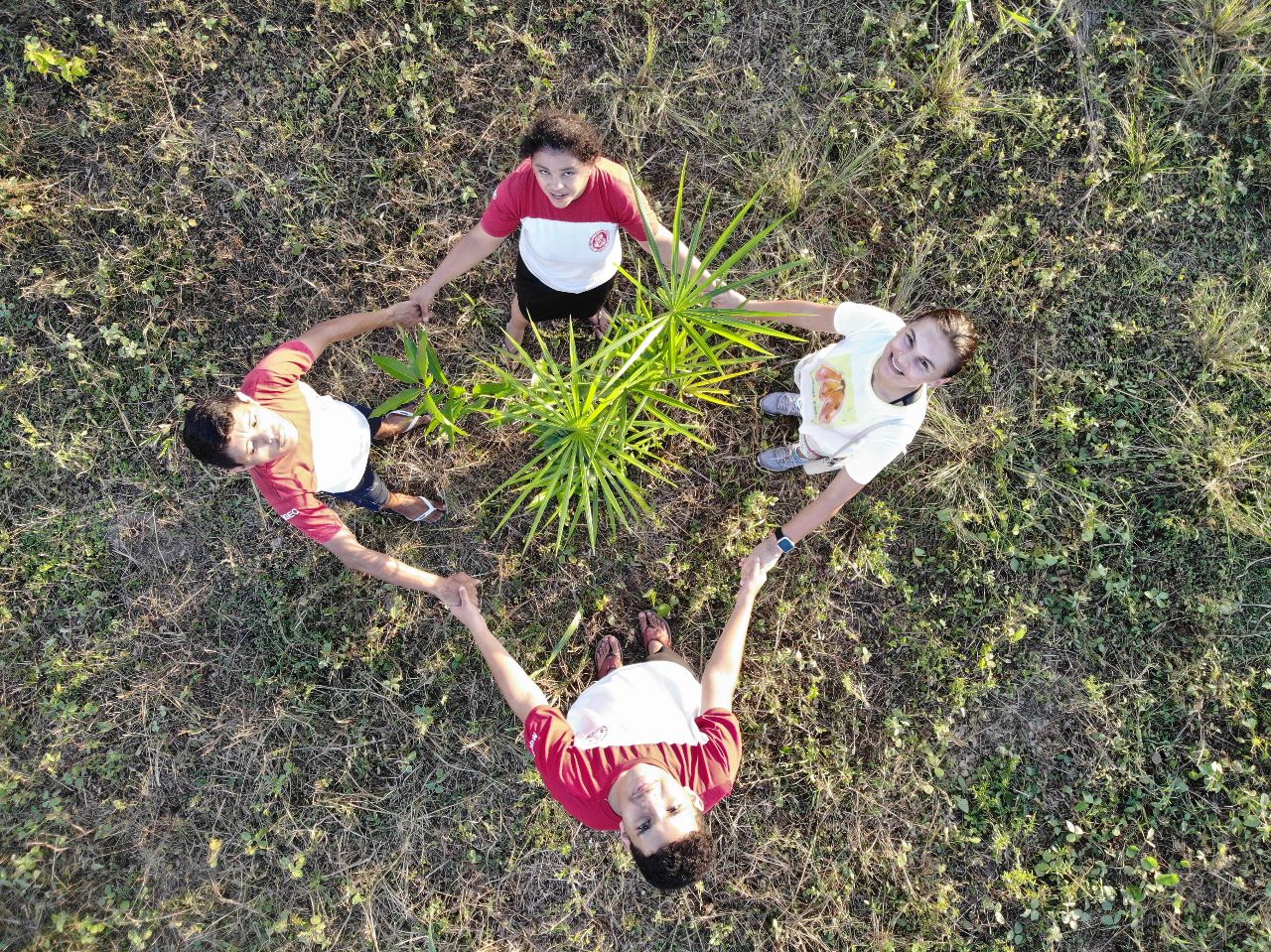 imagem mostra estudantes concorrentes a melhor escola do mundo em uma área de gramado, segurando as mãos em volta de uma muda plantada no chão.