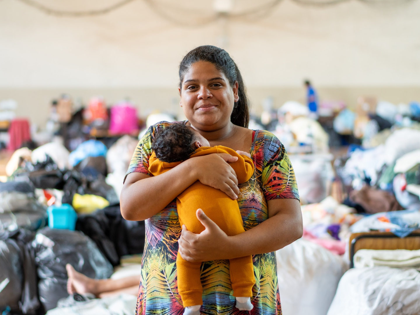 Imagem mostra uma mulher negra com roupa colorida segurando no colo um bebê recém-nascido coberto por uma manta. 