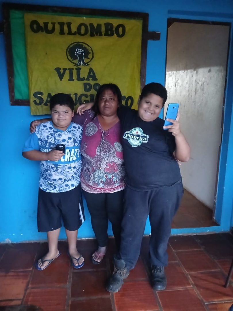 magem mostra uma mulher negra abraçando os filhos, dois meninos negros. Ao fundo, uma parede azul e um painel amarelo com o nome do quilombo: Vila do Sabugueiro.