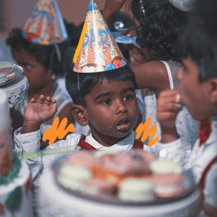 Imagem de capa para matéria sobre festas de aniversário pelo mundo mostra um menino negro usando chapéu de aniversário ao redor de uma mesa decorada de festa