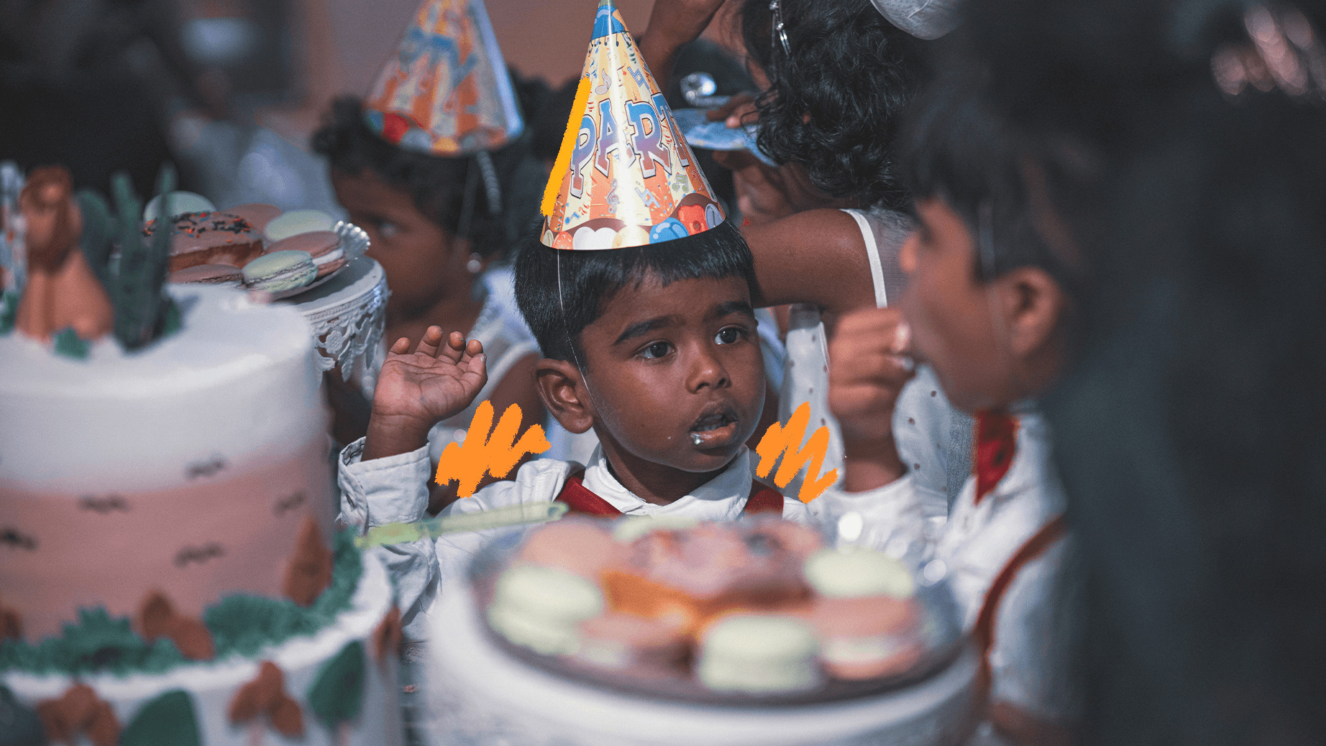 Imagem de capa para matéria sobre festas de aniversário pelo mundo mostra um menino negro usando chapéu de aniversário ao redor de uma mesa decorada de festa